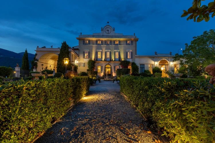 The villa's main façade lit at dusk, at the end of a hedge-lined gravel path